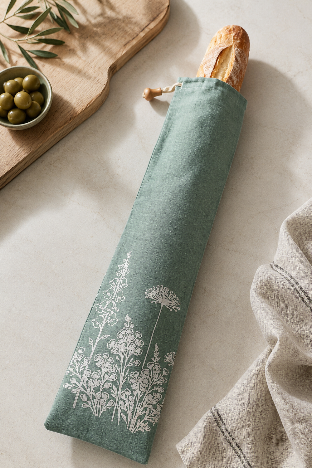 duck egg Green linen baguette bag with white floral design on a light surface with bread and olives.