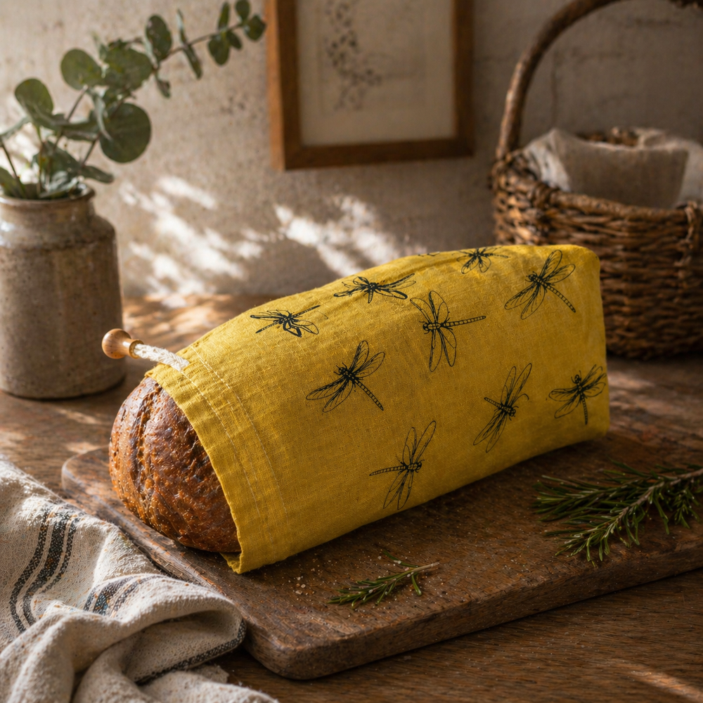 Loaf of bread wrapped in a yellow bread bag made from linen with black dragonfly patterns on a wooden board.
