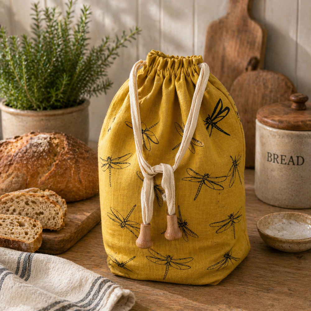 Yellow drawstring linen bread bag with dragonfly pattern on a wooden table with bread and kitchen items.