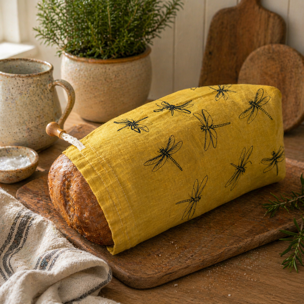 Loaf of bread wrapped in a yellow linen cloth bread bag with black dragonfly patterns on a wooden board.