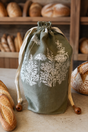 A sage green linen drawstring bread bag with white botanical illustrations, styled on a rustic wooden table with fresh bread loaves in the background.