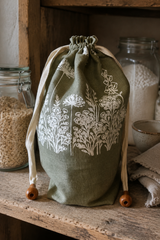 A sage green linen drawstring bread bag with white floral embroidery, displayed on a rustic wooden shelf next to glass jars containing grains.