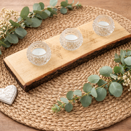 Decorative setting with wooden board, glass candle holders, and eucalyptus leaves on a woven mat.