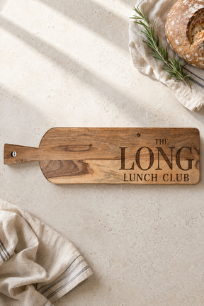 A wooden serving board with a handle and engraved text reading 'THE LONG LUNCH CLUB' lies on a light stone surface. The board is styled with a loaf of bread on a cloth in the top right and a folded napkin in the bottom left, with soft diagonal shadows across the scene.
