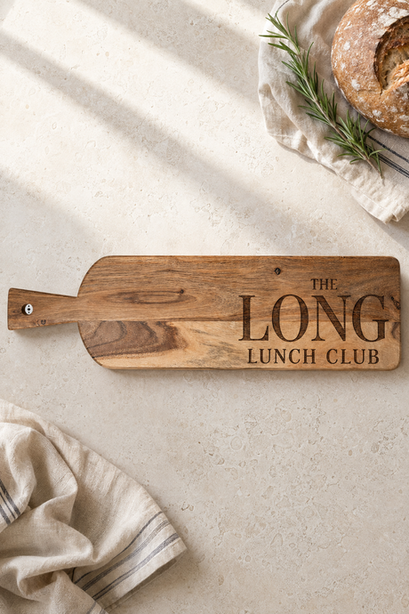 A wooden serving board with a handle and engraved text reading 'THE LONG LUNCH CLUB' lies on a light stone surface. The board is styled with a loaf of bread on a cloth in the top right and a folded napkin in the bottom left, with soft diagonal shadows across the scene.
