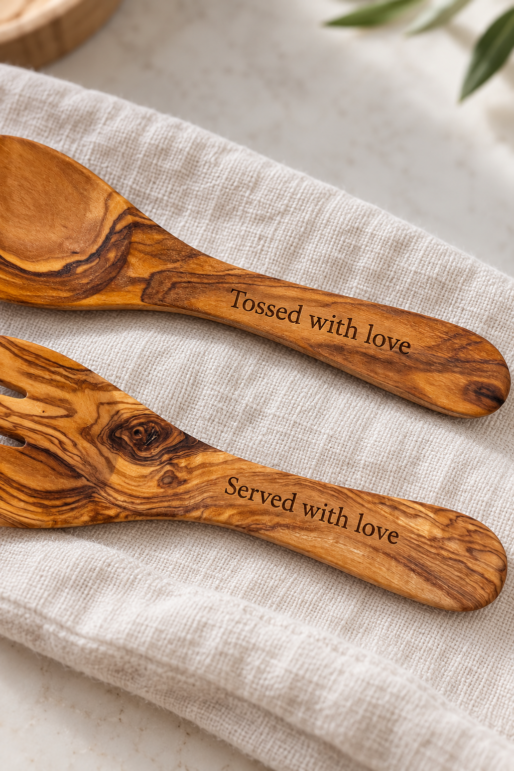 Close-up of olive wood handles engraved with “Tossed with love” and “Served with love”, showing the lettering and natural wood grain.