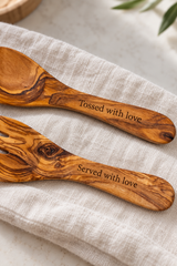 Close-up of olive wood handles engraved with “Tossed with love” and “Served with love”, showing the lettering and natural wood grain.