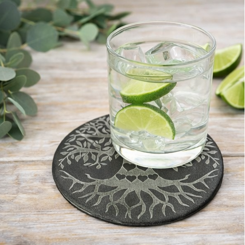 Set of four black coasters with leather  tree-of-life design on a wooden surface.