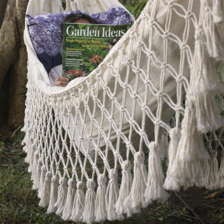 Macrame hammock with tassels holding a magazine titled 'Garden Ideas' outdoors.
