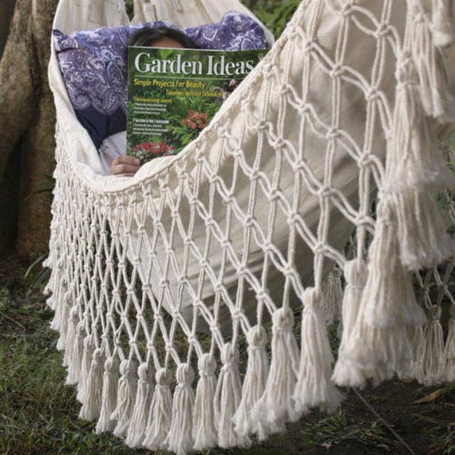 Macrame hammock with tassels holding a magazine titled 'Garden Ideas' outdoors.