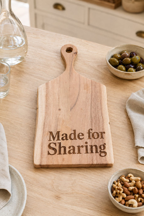 A rectangular wooden serving board with a handle and the engraved text 'Made for Sharing' sits on a light wood table, surrounded by dining props like bowls of olives and nuts, a glass, and a napkin.