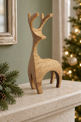A minimalist wooden reindeer figurine with natural grain stands on a stone mantelpiece, with a blurred Christmas tree and greenery in the background.