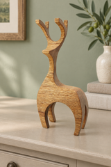A small sculptural deer figurine made of natural wood with visible grain, displayed on a light-colored cabinet in a styled interior setting with a vase and framed art in the background.