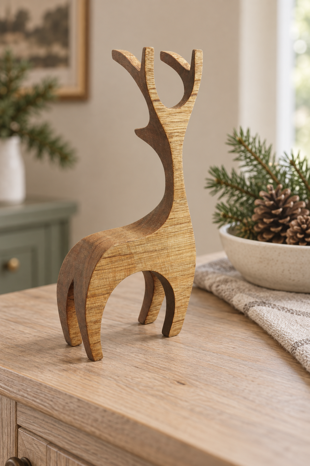 A minimalist wooden reindeer figurine with a natural wood grain finish, styled on a light wood console table next to a bowl of pinecones and evergreen branches.