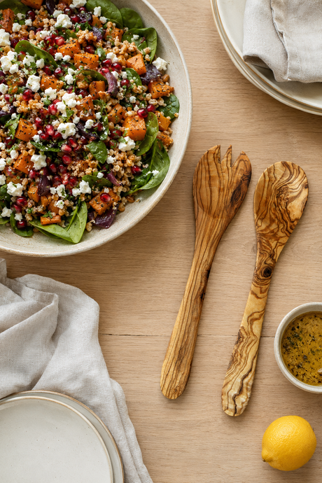 An overhead styled food scene on a light wood table showing a set of olive wood salad servers including a spoon and fork large bowl of salad with greens, crumbled cheese, and cubes of roasted sweet potato, alongside two wooden salad servers, a small bowl of dressing, a lemon, and folded napkins.