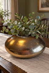 Antique gold pebble bowl with eucalyptus and olive foliage on a linen-covered wooden dining table.