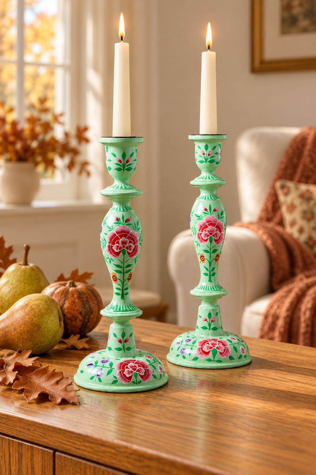 A pair of mint green ceramic candlestick holders with pink floral designs, holding white taper candles and set on a wooden table in a cozy living room.