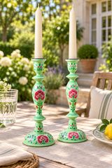 A pair of mint green metal candlestick holders with hand-painted pink floral motifs, holding white taper candles on an outdoor patio table.