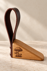 A wooden and leather door stopper with an engraved mushroom design on the base, photographed on a neutral textured beige surface.