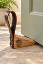 A wooden and leather door stopper resting on a stone floor next to a painted green door. The stopper features a triangular wooden base with a dark brown leather loop attached by metal rivets, and an engraved tree logo on the front.