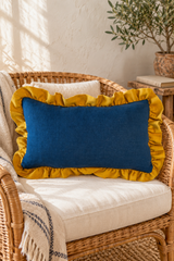 Plain rectangular blue linen cushion with mustard ruffle on a wicker chair beside an olive tree..