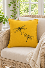 A square yellow linen pillow with black dragonfly, resting on a beige cushion of a wicker armchair in a bright, sunlit room.