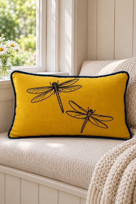 Rectangular mustard linen cushion with two black dragonflies and deep blue piping on a cream window seat beside daisies.
