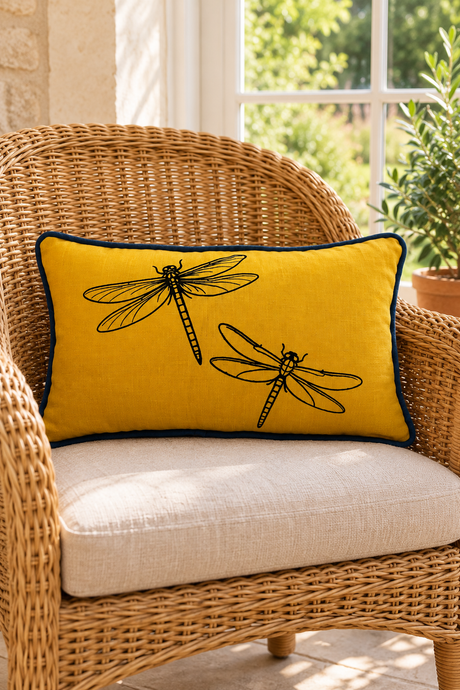 Rectangular mustard cushion with black dragonfly print and navy blue piping on a wicker armchair beside a sunny window.