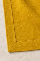 Reverse corner of a mustard linen napkin showing the diagonal mitred join, double-fold hem and yellow stitching.