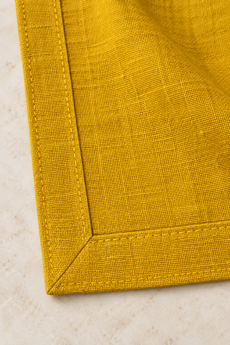 Reverse corner of a mustard linen napkin showing the diagonal mitred join, double-fold hem and yellow stitching.