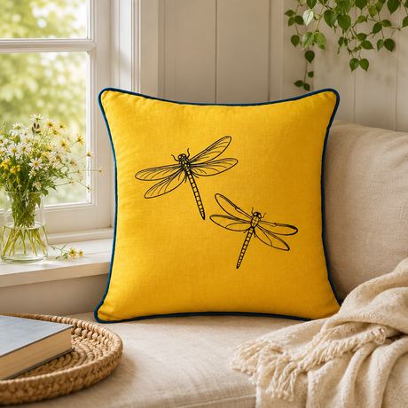 Mustard linen cushion cover with black dragonfly print and deep blue piping styled on a cream window seat.