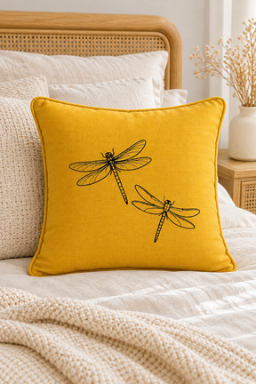 A square yellow linen pillow with two black  dragonflies, styled on a bed with neutral-toned bedding and a wooden headboard.