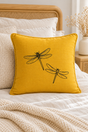 A square yellow linen pillow with two black  dragonflies, styled on a bed with neutral-toned bedding and a wooden headboard.
