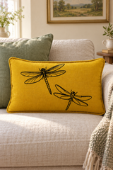 Rectangular mustard linen cushion with two black dragonflies and matching piping on a cream sofa beside a sage cushion.