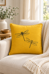 A square yellow Linen pillow with black dragonfly, resting on a light beige armchair in a cozy living room setting with a cream knit blanket.