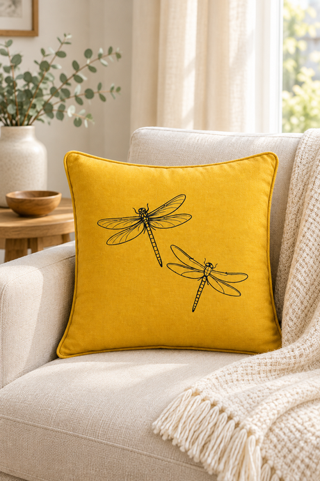 A square yellow Linen pillow with black dragonfly, resting on a light beige armchair in a cozy living room setting with a cream knit blanket.