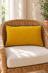 Plain mustard rectangular cushion with deep blue piping on a wicker armchair with a cream seat pad.