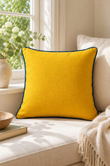 Square mustard linen cushion with deep blue piping on a cream window seat beside eucalyptus and a woven throw.