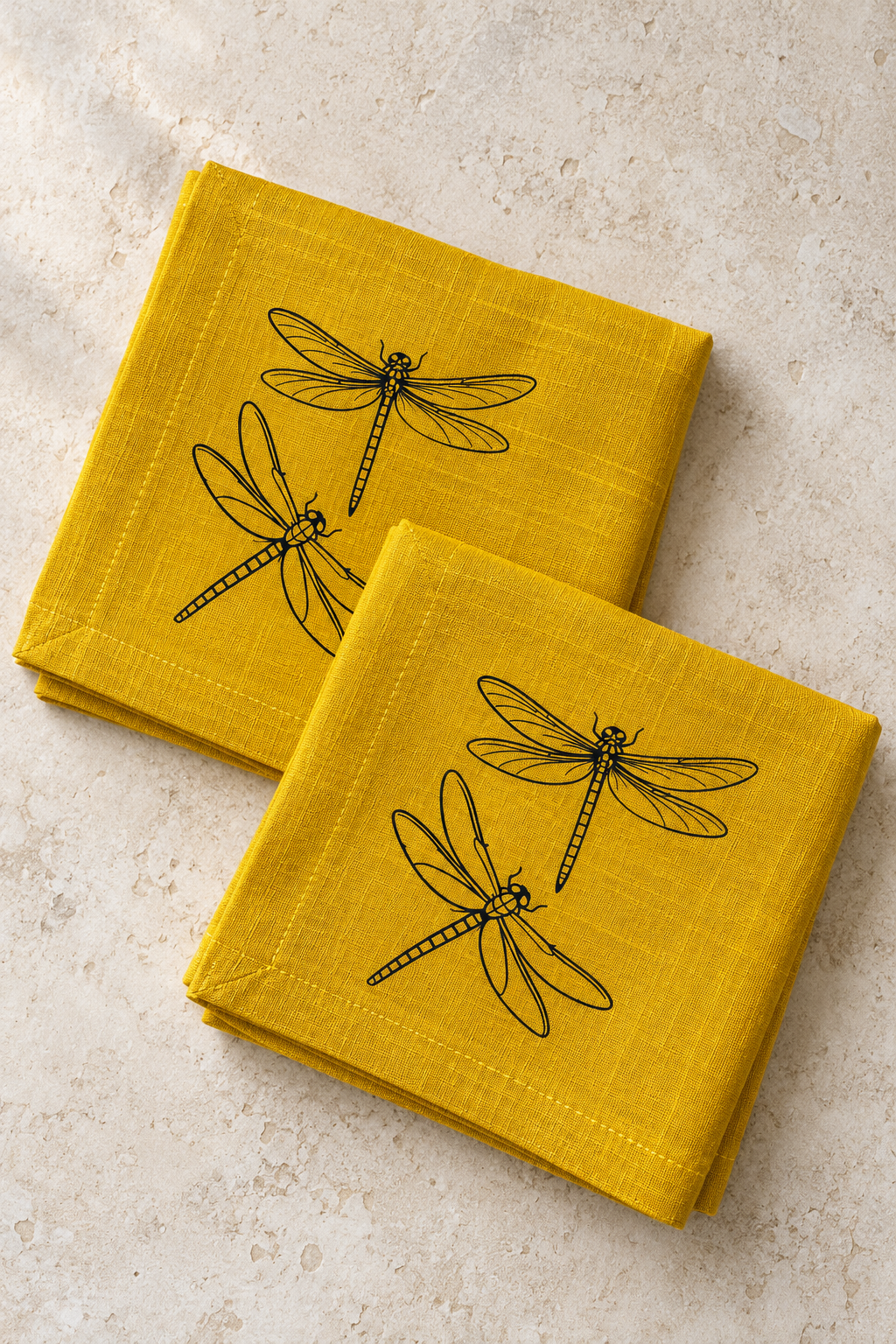Two folded mustard yellow linen napkins with black dragonfly prints and matching stitched hems on pale stone.