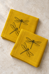 Two folded mustard yellow linen napkins with black dragonfly prints and matching stitched hems on pale stone.