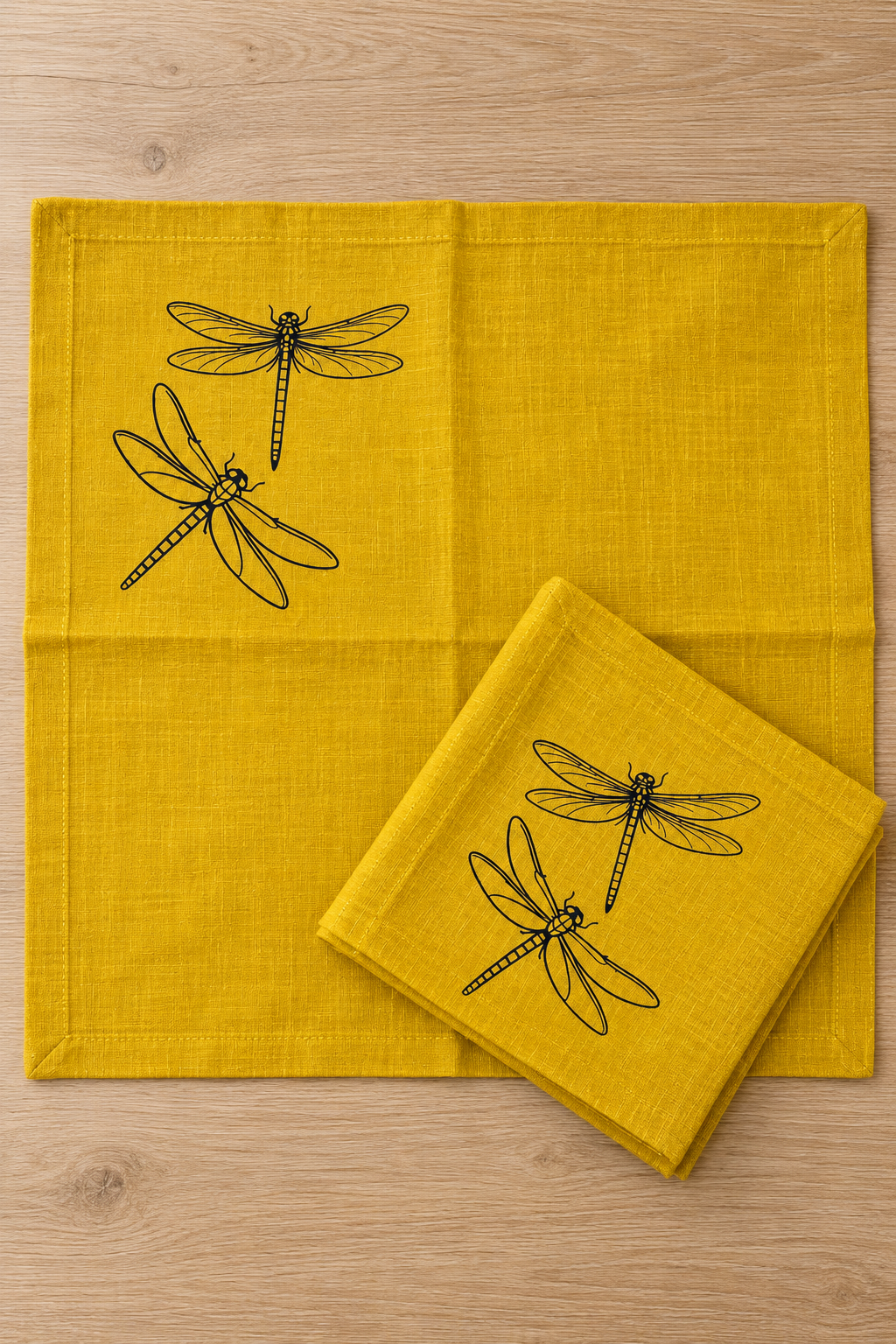 Mustard linen napkin opened flat beside a folded matching napkin, showing the black dragonfly print near one corner.