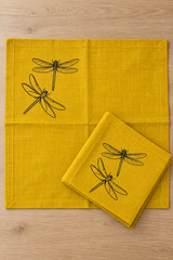 Mustard linen napkin opened flat beside a folded matching napkin, showing the black dragonfly print near one corner.