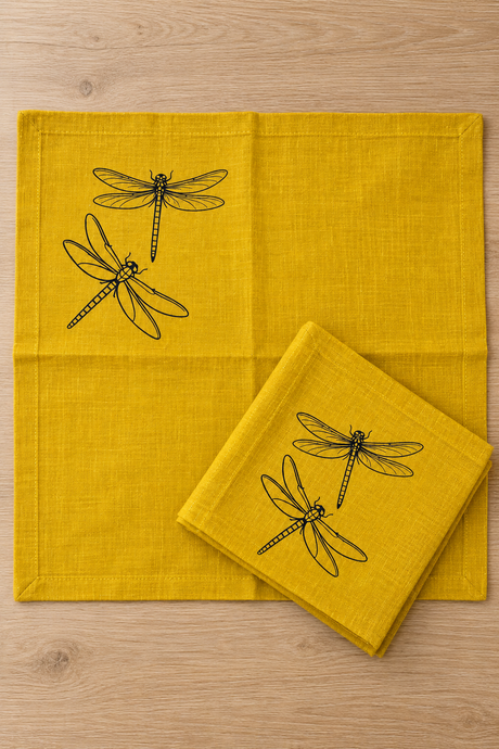 Mustard linen napkin opened flat beside a folded matching napkin, showing the black dragonfly print near one corner.