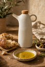 A matte ceramic olive oil dispenser with a cork stopper stands on a rustic wooden table beside a small bowl of olive oil, bread, olives, and rosemary in a warm, styled kitchen setting.