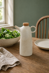 A matte off-white ceramic oil bottle with a cork stopper and curved handle sits on a wooden dining table, styled with a bowl of salad, a plate, and a linen napkin in a softly lit room.