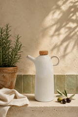 A matte white ceramic oil bottle with a cork stopper and loop handle sits on a stone ledge beside a potted rosemary plant, a folded cream cloth, and a small olive branch against a textured beige wall with leaf shadows.