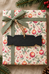 A wrapped gift box with festive Christmas paper featuring red ornaments and green holly on a cream background, tied with an olive green ribbon and bow, and topped with a Slate Christmas Cracker-shaped Gift tag.