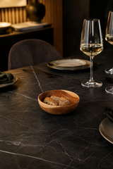 A small round wooden bowl filled with bread sits on a dark marble dining table set with plates, cutlery, and wine glasses in a dimly lit restaurant or dining room.