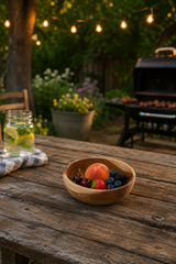 A rustic wooden bowl filled with fresh fruit sits on a weathered wooden table in a backyard setting with a grill, string lights, and a mason jar beverage nearby.
