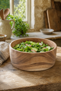 A round wooden salad bowl filled with fresh green salad, set on a rustic wooden table in a warm, sunlit kitchen setting.